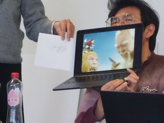 Two people posing with a drawing and a laptop showing a funnyy meme with the face of an elder sage.